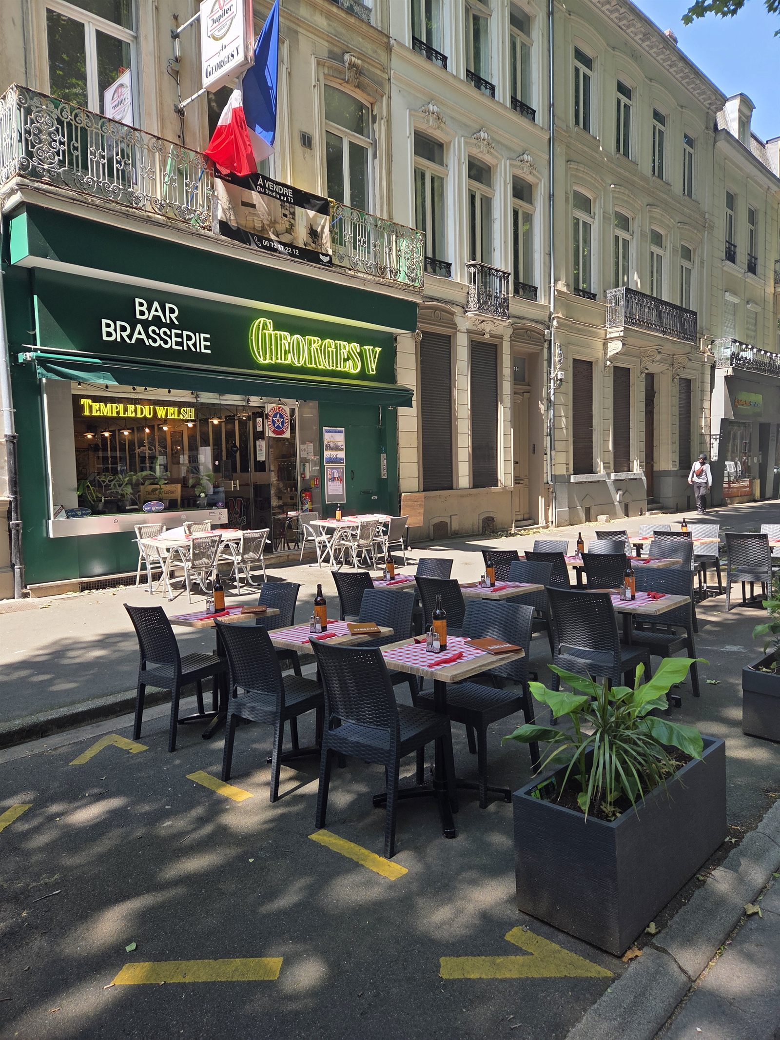 Façade du Georges V en plein soleil avec terrasse aux nappes vichy, drapeau français et enseigne Temple du Welsh, 104b Boulevard de la Liberté à Lille